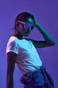 Stylish brazilian female in fashionable sunglasses and outfit touching hair while standing on purple background in studio with fluorescent light