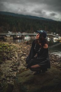 Young woman crouching on rock at riverbank against cloudy sky