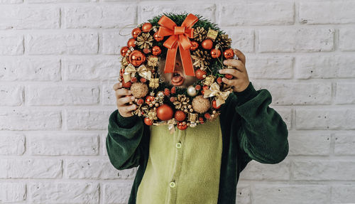 Little boy with a christmas wreath grimaces, shows his tongue against a white brick wall