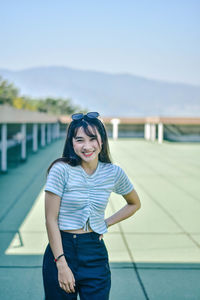 Portrait of young woman smiling while standing on floor at building terrace against sky