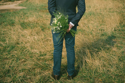 Low section of bridegroom holding bouquet while standing on grassy field in park