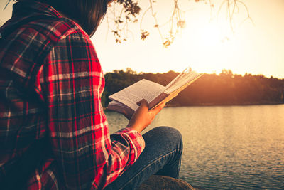 Man holding book while sitting by lake against sky during sunset