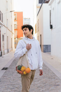 Side view of content male standing with eco friendly mesh bag full of fresh fruits in city street looking away