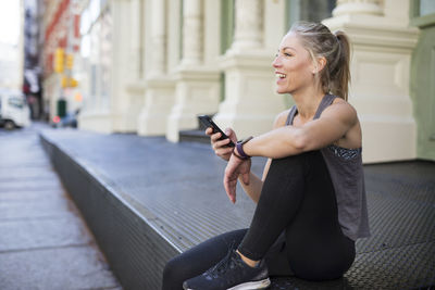 Happy athlete using mobile phone while sitting at sidewalk in city