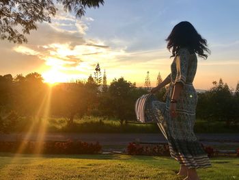 Side view of young woman standing on field against sky during sunset