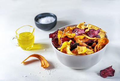 Healthy mixed vegetable chips in a bowl on light surface with marine salt and oil