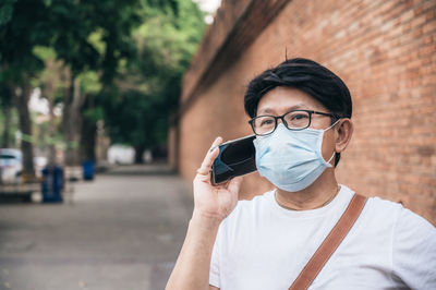 Man wearing mask talking over smart phone while standing against brick wall