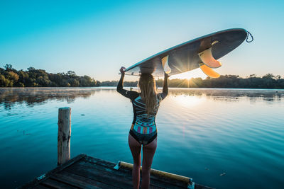 Rear view of woman with surfboard standing on pier over lake against blue sky