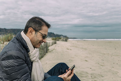 Man using phone while sitting at beach against sky