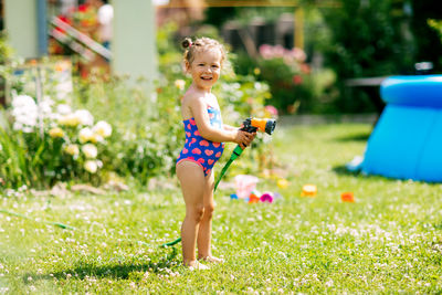 Charming baby girl waters plants with a hose in the garden in the backyard of the house on a sunny 