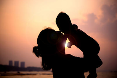 Silhouette woman playing with son at beach against sky during sunset