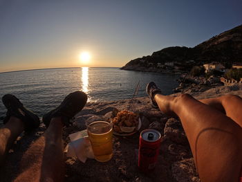 Low section of man sitting at beach against sky during sunset