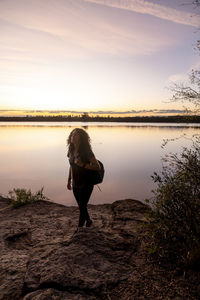 Rear view of woman standing by lake against sky during sunset