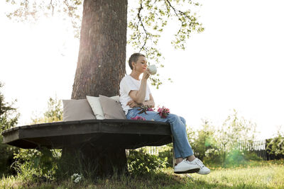 Smiling woman holding coffee cup sitting on bench around tree trunk in garden