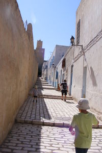 Rear view of man and boys walking in alley amidst buildings on sunny day