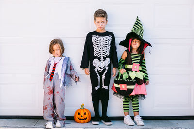 Full body of group of little kids dressed in various halloween costumes with carved jack o lantern standing near white wall on street