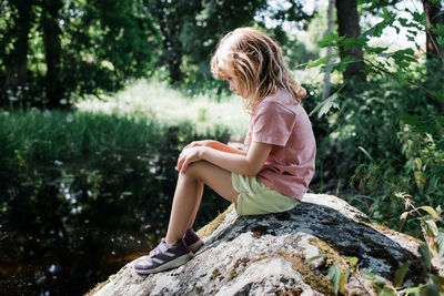 Young girl looking sad and alone on a rock by a river in summer