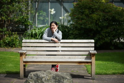 Portrait of smiling young woman standing by wooden bench at park