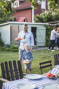 Father and son walking with food plate towards dining table for garden party in backyard during weekend
