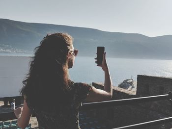 Rear view of woman taking selfie while standing against sea and sky