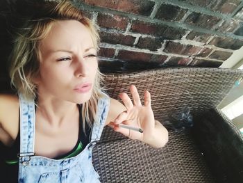 High angle view of woman smoking cigarette while sitting on wicker furniture against brick wall