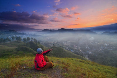 Man sitting on grassy field and pointing at mountain range during sunset