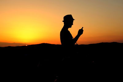 Silhouette man holding mobile phone while standing against sky during sunset