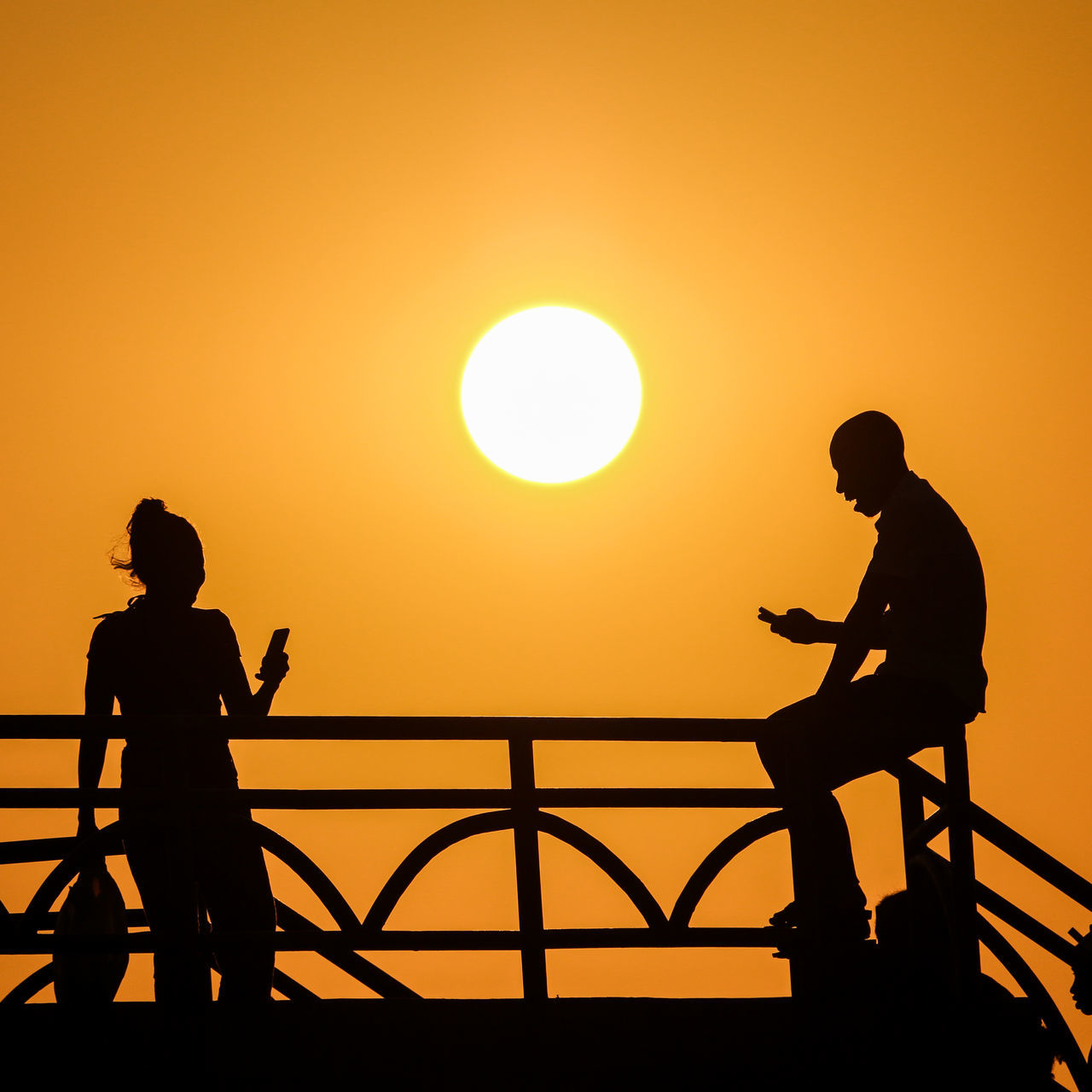 sunset, silhouette, real people, orange color, lifestyles, men, leisure activity, sky, beauty in nature, sun, standing, people, scenics - nature, two people, nature, water, togetherness, railing, clear sky, outdoors