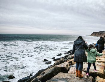 Mother with daughter walking on rocky sea shore against sky