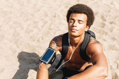 Shirtless male athlete listening to music through in-ear headphones while sitting at beach