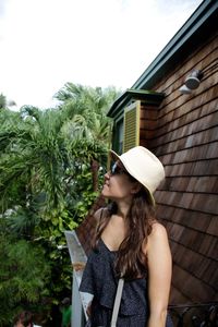 Woman in sunglasses and hat standing by house against sky