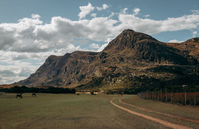 Trail and fences leading into a mountain with a blue cloudy sky.