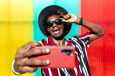 Optimistic young tattooed african american male in bright striped shirt and hat with trendy sunglasses and wristwatch taking selfie on mobile phone while standing against colorful wall