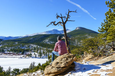 Rear view of man sitting on rock against mountains during winter