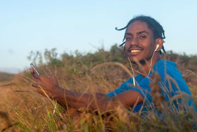 Portrait of young man with sitting on field holding a mobile phone against sky