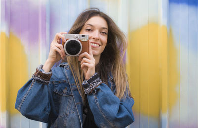 Portrait of smiling woman holding camera against curtain