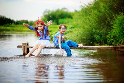 Portrait of cheerful siblings sitting on wood over lake