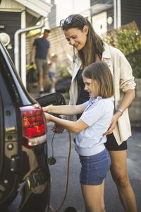 Portrait of mother, father and two daughters standing by car at electric vehicle charging station