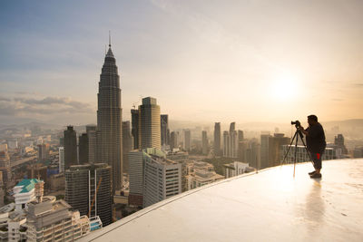 Full length of man photographing skyscrapers on building terrace using camera against sky