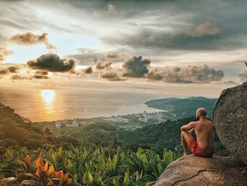 Shirtless man looking at sea while sitting on cliff against sky during sunset