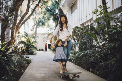 Smiling daughter skateboarding with help of mother on footpath