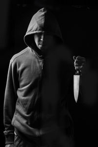 Man wearing hooded jacket holding knife while standing against black background