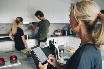 Mature woman with technologies looking at man and daughter standing in kitchen