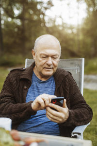 Senior gay man using smart phone sitting on chair at yard