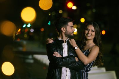 Portrait of smiling girlfriend holding boyfriend while standing in illuminated city at night
