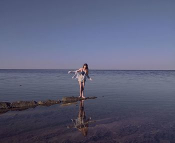 Full length young beautiful woman standing on beach against clear sky