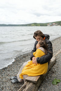 Side view of mother and daughter sitting hugging and kissing on the sea shore enjoying a winter day