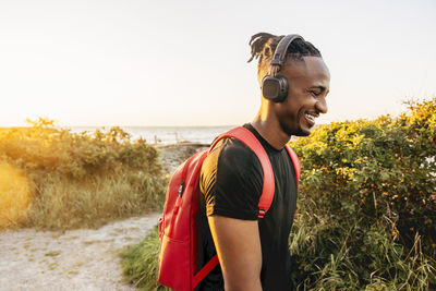 Smiling young man wearing backpack and wireless headphones against sky