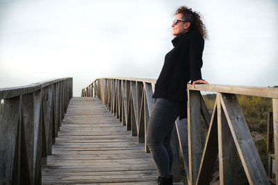 Side view of woman wearing sunglasses while standing on footbridge against sky