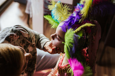 From above cropped unrecognizable woman applying makeup on cute little girl in colorful feather headgear costume during carnival preparation at home